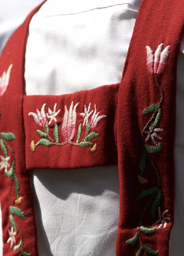 Close-up of traditional costume with red embroidered shoulder strap, worn by a person in white. | © Kleinwalsertal Tourismus eGen • Photographer: Hans Wiesenhofer