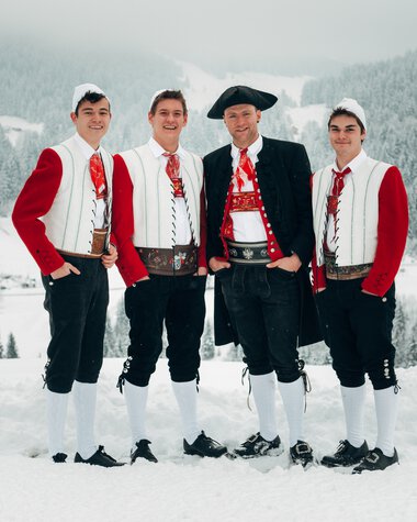 Four young men in traditional costumes standing in snowy mountain landscape, smiling and posing together in winter. | ©   Kleinwalsertal Tourismus eGen • Photographer: Stefan Klauser