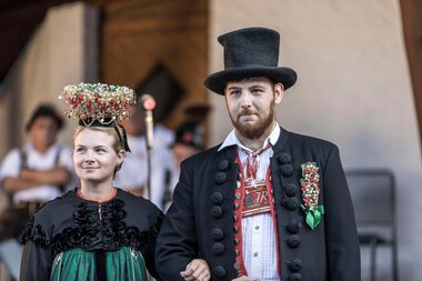 A man and a woman in traditional costumes at an alphorn festival, with the woman wearing a floral crown on her head. | © Kleinwalsertal Tourismus eGen • Photographer: Frank Drechsel