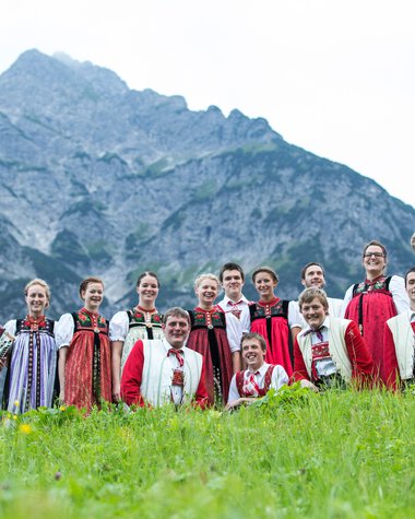 Group of people in traditional clothing in front of a mountain backdrop, some with musical instruments, standing on the grass. | ©  Kleinwalsertal Tourismus eGen • Photographer: Frank Drechsel