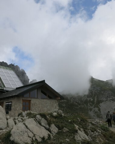 Berglandschaft mit Wolken, ein kleines Haus, große Steine und eine Gruppe von Menschen auf einem Wanderweg | © Kleinwalsertal Tourismus eGen • Fotograf: Antje Pabst