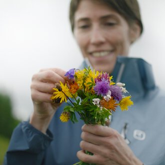 Frau beim Pflücken eines bunten Blumenstraußes im Freien, mit einem Lächeln auf dem Gesicht. | © Kleinwalsertal Tourismus eGen • Fotograf: Clemens Paul