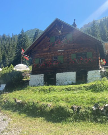 Gemütliches altes Chalet aus Holz mit bunten Fensterläden und Blumen, in einer bergigen Landschaft bei Sonnenschein. | © Kleinwalsertal Tourismus eGen