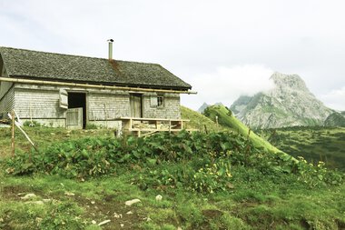 Ein ländliches Haus im Gebirge mit grünen Wiesen, Berge im Hintergrund und bewölktem Himmel. | © Kleinwalsertal Tourismus eGen