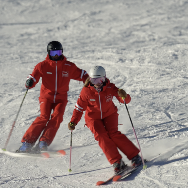 Two skiers in red suits during a ski lesson on snow, one with a helmet and the other with a cap, skiing on a snowy slope. | © Kleinwalsertal Tourismus | Skischule Hirschegg