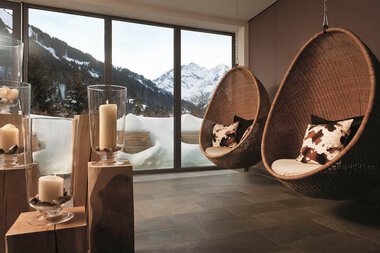 Gemütlicher Raum im Ifen Travelcharme Hotel mit Blick auf verschneite Berge durch große Fenster, zwei Hängesessel und Kerzen. | © Arosa Ifen Hotel
