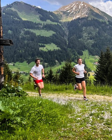 Two people running on a trail in the mountains with green meadows and high peaks in the background. | © Kleinwalsertal Tourismus eGen 