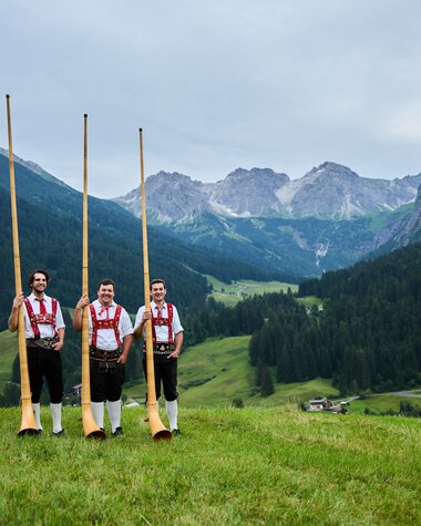 Drei Männer in Trachten stehen in einer grünen Wiese mit Alphörnern, umgeben von Bergen und einer bewaldeten Talregion. | © Kleinwalsertal Tourismus eGen • Fotograf: Oliver Farys