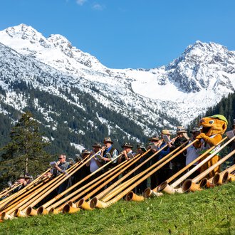 Gruppenfoto beim Alphornfestival 2024 mit Musikern in traditioneller Kleidung und Alphörnern, vor atemberaubender Bergkulisse mit schneebedeckten Gipfeln. | © Kleinwalsertal Tourismus eGen • Fotograf: Frank Drechsel