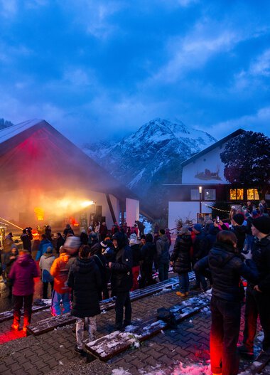 People gather at the Alphorn Festival 2024 in a mountain area at dusk, with an outdoor stage and snow-capped peaks in the background. | © Kleinwalsertal Tourismus eGen • Photographer: Frank Drechsel